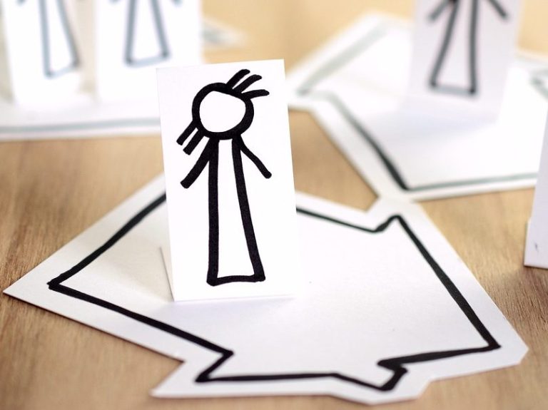 Zwei weiße Papierschnitte mit einfachen Strichfiguren und Pfeilen auf einem Tisch.